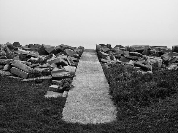 Black and white landscape photograph of a sidewalk leading to nowhere, but it actually goes to the shore of Lake Michigan, which is hidden in a thick blanket of early morning fog. 