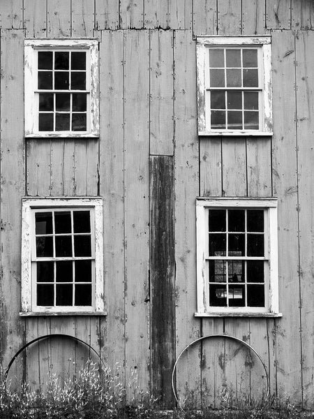 Black and white photograph of a one wall of a big prairie farmhouse with four white-framed windows and two leaning metal wheel rims