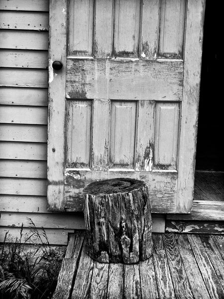 Black and white photograph of an old tree stump holding open the door of an old midwestern farmhouse.