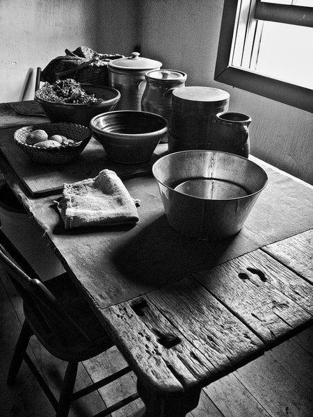 Black and white photograph of a rough and rustic kitchen table covered with jars and bowls, found in an old farmhouse. Imagine the stories this old table could tell.