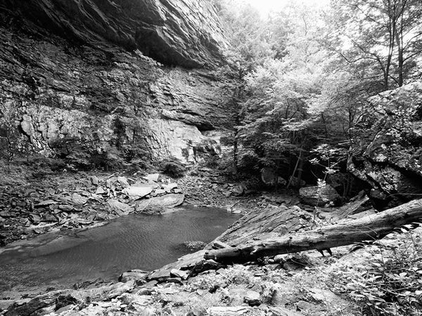Black and white landscape photograph of the rocky pool at the base of Ozone Falls, Tennessee. The pool is fed by Fall Creek, which falls 110' from the cliff above, and flows out through a subterranean passage and back above ground further downstream.