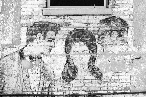 Black and white photograph of an old, fading, and peeling fashion ad on a brick wall in downtown Milwaukee, showing a painting of people in 60s-style evening wear.