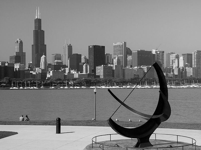Black and white photograph of the great Chicago skyline, as seen from the Adler Planetarium location on the lakeshore of Lake Michigan. In the foreground is "Man Enters the Cosmos" sculpture by Henry Moore.