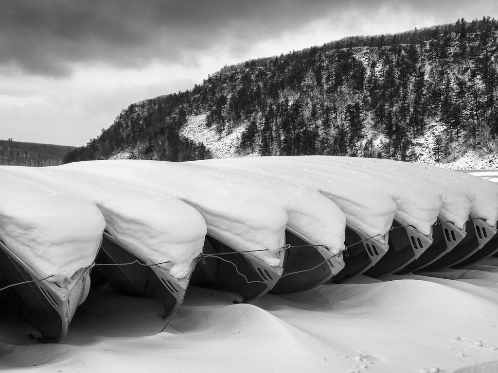 Black and white landscape photograph of row boats stored for winter, covered in a blanket of snow at Devil's Lake in Wisconsin. Devil's Lake was formed naturally by the action of glaciers during the last ice age.