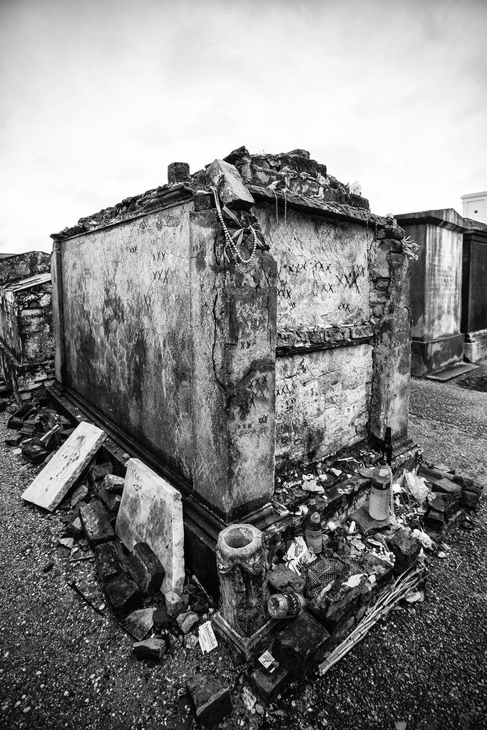 Black and white fine art photograph of a voodoo tomb in New Orleans' famous St. Louis Cemetery No. 1. The old tomb is marked by characteristic triple Xs and littered with offerings around its base. 