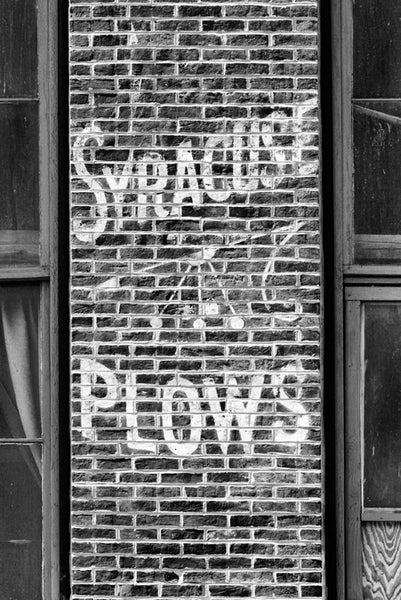 Black and white photograph of a vintage hand-painted advertisement for Syracuse Plows, found on a brick wall near the waterfront in downtown Nashville, Tennessee.