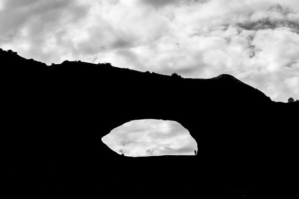 Black and white landscape photograph of one of the many arches to be found in the desert landscape of Utah. In this silhouette, two small figures can be seen inside the eye of the arch.