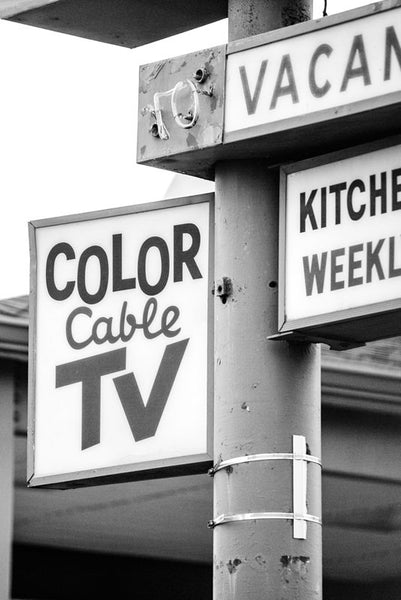 Black and white photograph of an old 60s style roadside motel sign for a motel that has since been demolished. The signs promise amenities like "Color Cable TV," and "Kitchenettes," and "Weekly Rates."