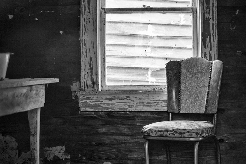 Black and white photograph of an old wooden table and a chair inside the home of the late blues musician Sleepy John Estes in Brownsville, Tennessee. The small shack where Estes lived has been preserved and relocated to a visitors center near the schoolhouse of Tina Turner, who also hails from Brownsville. The wall of Turner's school is visible through the window.