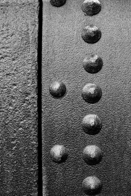 Black and white industrial detail photograph of rusty rivets on a curved sheet of rusty metal, part of an antique machine.