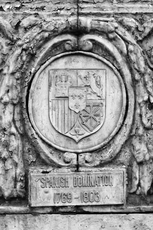 Black and white photograph of an ornate street pole crest, part of a series illustrating the nations that have ruled over Louisiana in the past, seen along Basin Street in New Orleans. The metallic sign says, "Spanish Domination 1769 - 1803." 