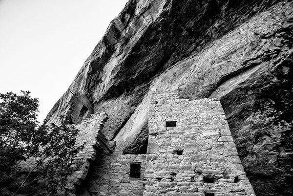 Black and white fine art photograph looking up at the tall vertical walls of Spruce Tree House, seen in its sheltered location under the overhanging edge of Mesa Verde in Colorado.