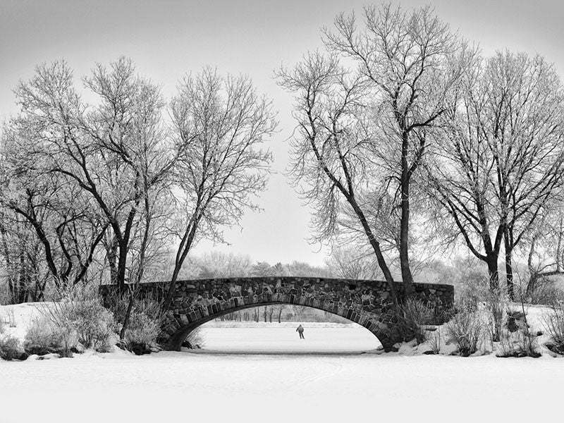 Black and white photograph of an ice skating pond with an old stone bridge arching over it. In the distance, the ice tester skates alone on the ice, ensuring that it's safe for the general public to skate.