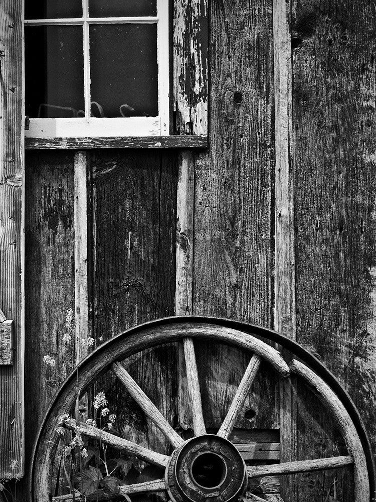 Black and white photograph of a broken wagon wheel leaned against the side of a textured wooden workshop.