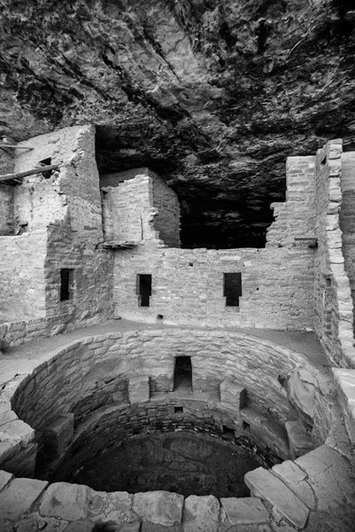 Black and white fine art photograph of ancient rooms and an open kiva at Mesa Verde, Colorado. The blackened streaks on the ceilings are the carbon remains of smoke from ancient cooking fires.