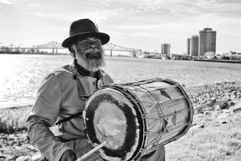 Black and white fine art photograph of a New Orleans street musician on the levee playing a drum and singing "When the saints go marching in." In the background is the Greater New Orleans Bridge (aka Crescent City Connection) over the Mississippi River.