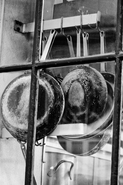 Black and white fine art photograph of pots and pans hanging in the window of a French Quarter gourmet restaurant kitchen in New Orleans, Louisiana. 