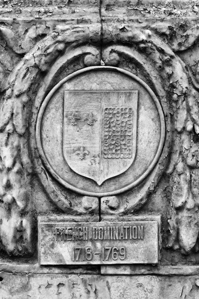 Black and white photograph of an ornate street pole crest, part of a series illustrating the nations that have ruled over Louisiana in the past, seen along Basin Street in New Orleans. The metallic sign says, "French Domination 1718 - 1769."