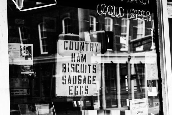 Black and white fine art photograph of store window on Front Street in downtown Memphis. The homemade sign offers Country Ham, Biscuits, Sausage, Eggs, and also Cold Beer.