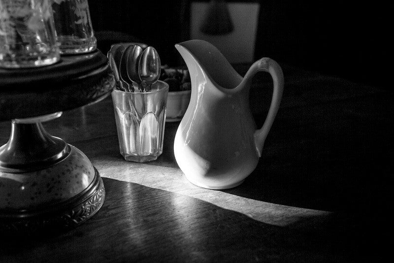 Black and white photograph of a shaft of morning sunshine shining over a dining table with a small cream pitcher in one of the 19th century houses at the historic South Union Shaker Village in Kentucky.
